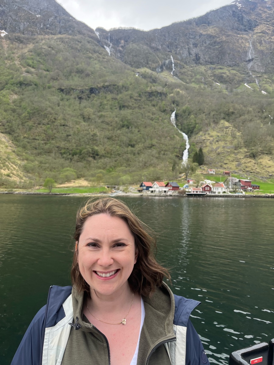 Hailly smiling on a boat in a fjord, with green mountains, a waterfall and red-roofed houses behind